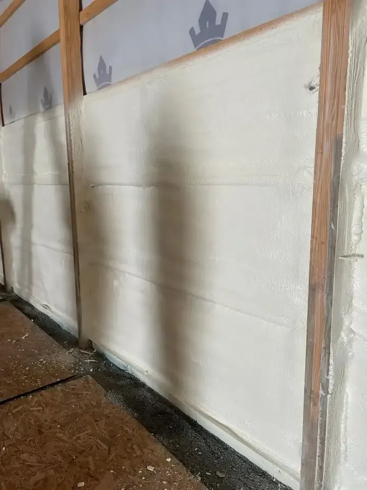 Spray foam insulation on pole barn walls between wood posts, with house wrap still exposed above the foam line