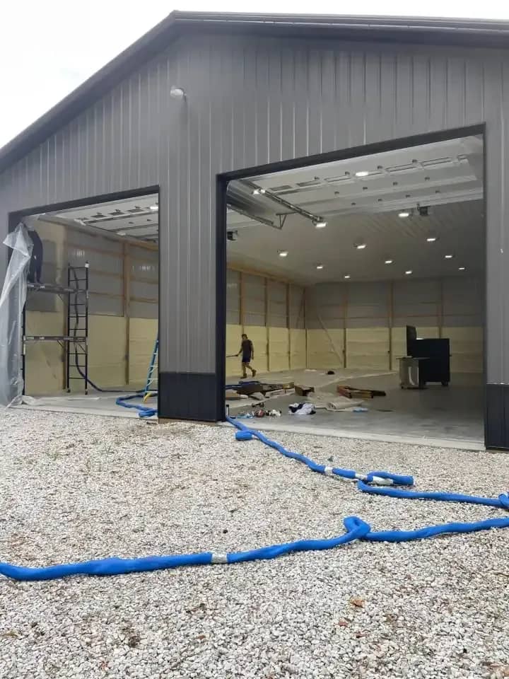 New gray metal pole barn with overhead doors open during a spray foam insulation job, spray hose on the gravel